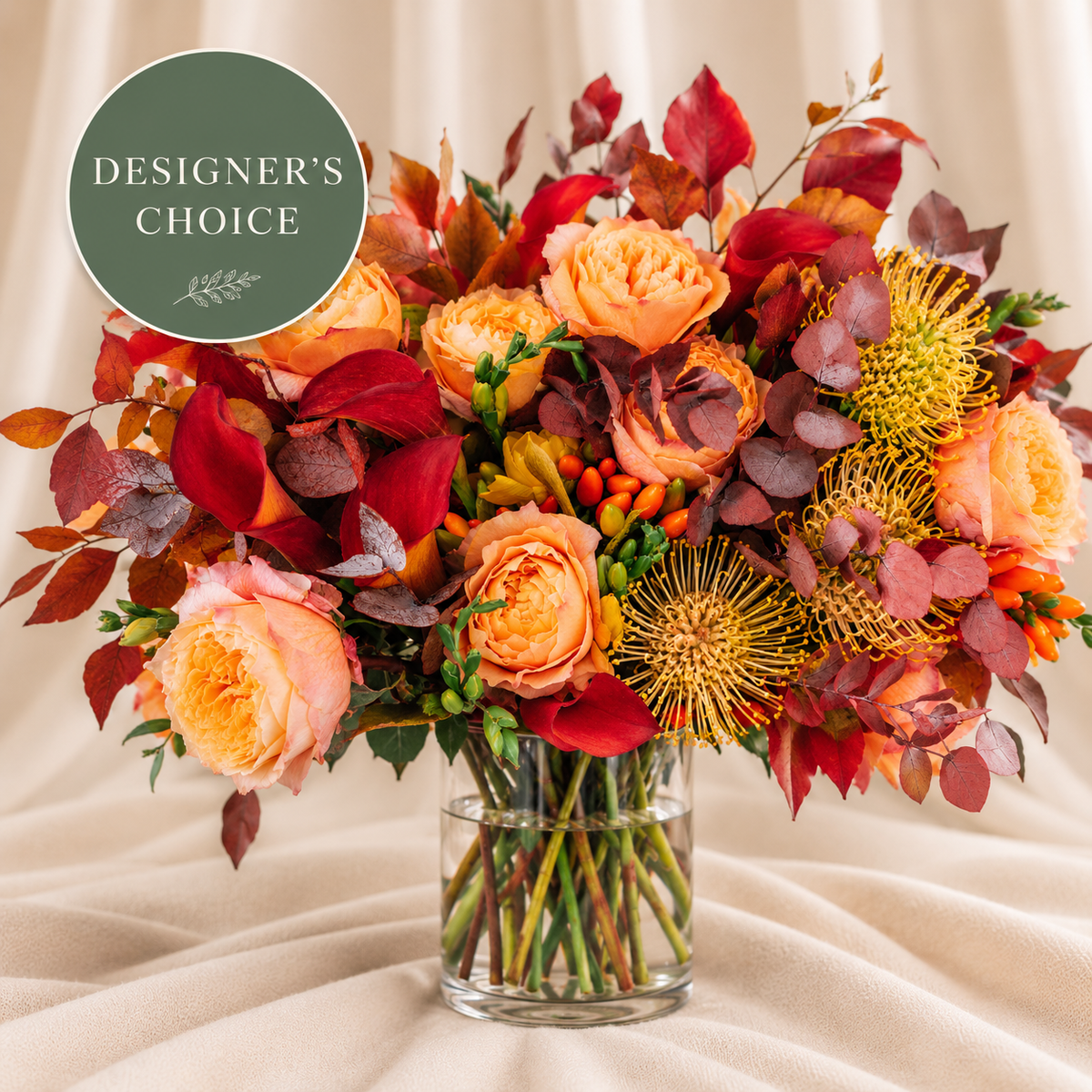 Designer’s Choice Fall Colors: peach roses, burgundy calla lilies, autumn leaves, berries, and golden protea in a clear glass vase